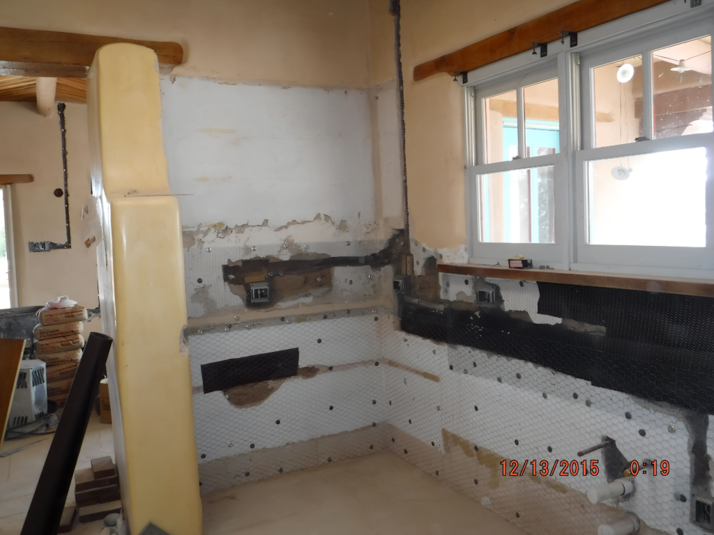 The image shows a kitchen under renovation, with exposed walls, a partially demolished section, and visible wiring and plumbing. There are also materials stacked nearby.