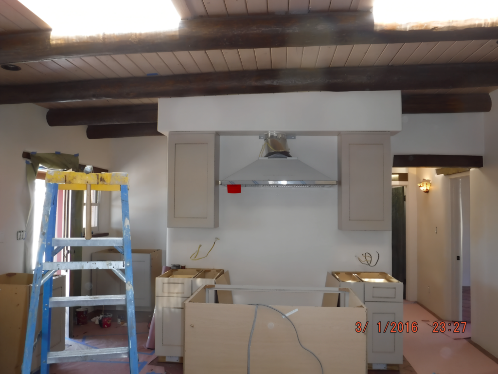 A kitchen under renovation featuring exposed wooden beams on the ceiling. Cabinets and a range hood are installed, while unfinished counters and a ladder indicate ongoing work. The space is being prepared for further updates and finishing touches.