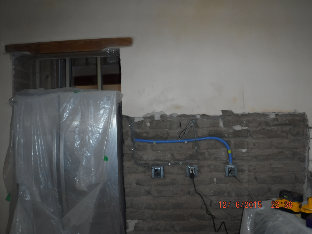 A kitchen wall in the process of renovation, with exposed electrical wiring and outlets. The surrounding area is partially covered with plastic sheeting for protection, and unfinished brickwork is visible, indicating ongoing construction work.