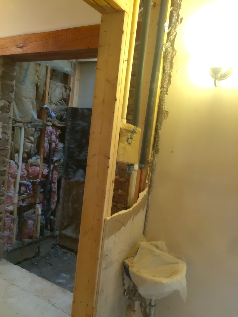 Another angle of the in-progress bathroom renovation, showing exposed pipes, insulation, and part of the sink wrapped in plastic. The room is under construction.