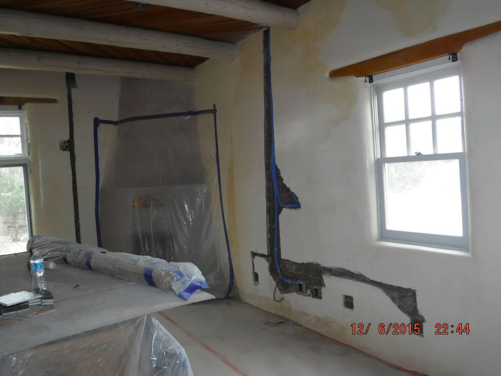 A bedroom in the middle of renovation, with exposed wiring and partially patched walls. Plastic sheets cover areas to protect from dust, and materials are rolled on the floor. Natural light streams through the windows, highlighting the construction progress.