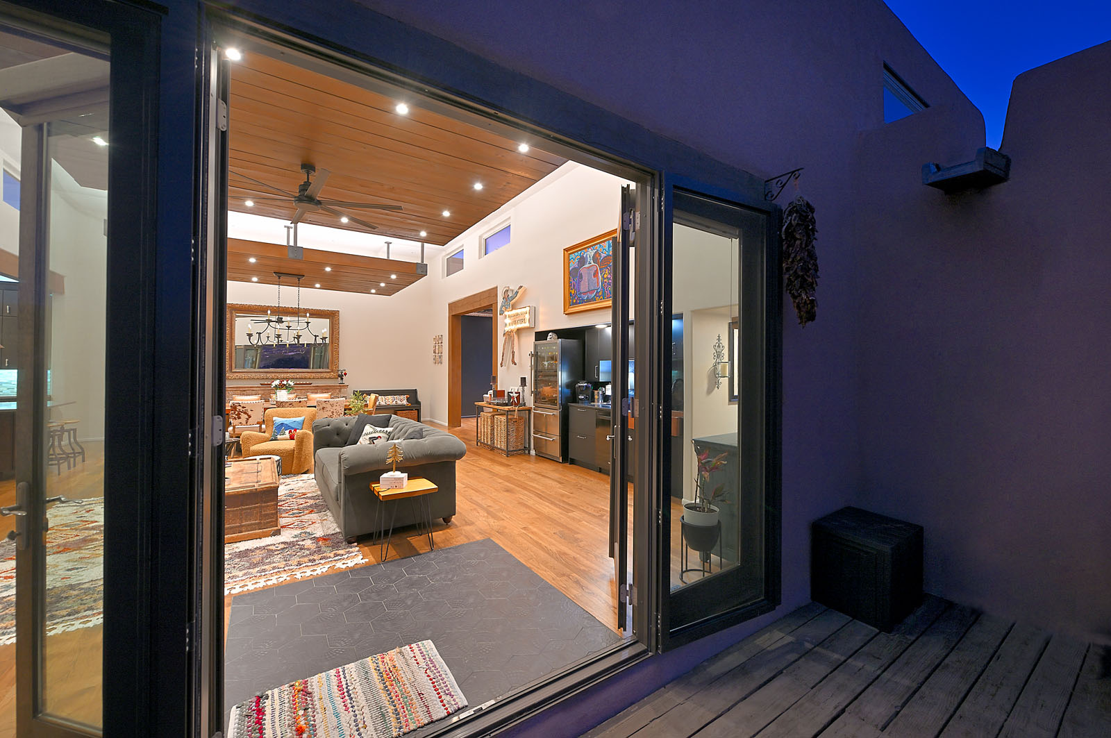 Evening view through large glass doors into a warmly lit and inviting Santa Fe home, showcasing a modern interior with wooden accents and comfortable furnishings.